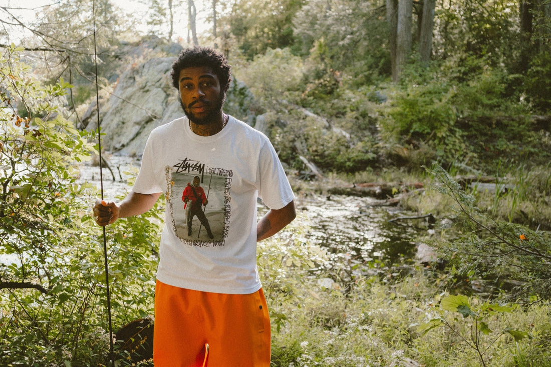 Stüssy x Denim Tears FW26 lookbook, orange fishing slicker pant by a stream in New York (photo @denimtears)